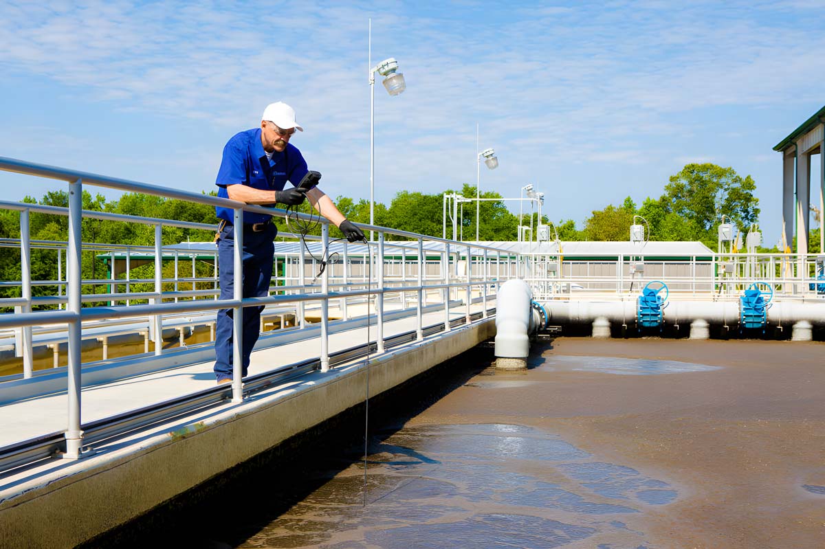 A wastewater operator standing on an open air causeway using scientific equipment to analyze the water quality in the tank below